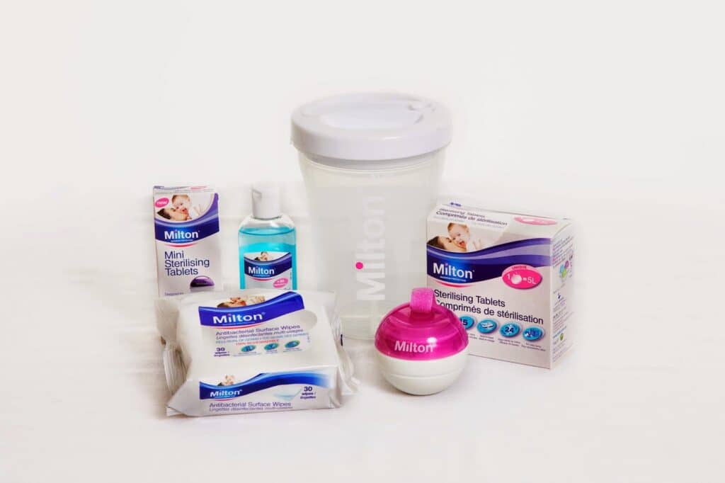A group of Milton-branded baby sterilising products, including sterilising tablets, antibacterial surface wipes, sterilising fluid, and a white container with a transparent lid, all arranged on a white surface with a white background.