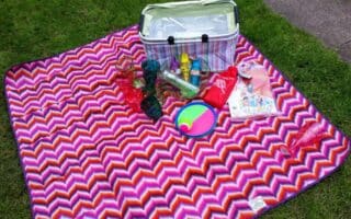 A colourful chevron-patterned picnic blanket on grass holds a striped cool box, plastic cups, suncream, insect repellent, a multicolour ball, a frisbee, a bag of snacks, and a children’s book, all ready for a picnic.