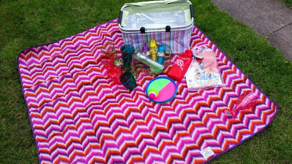 A colourful zigzag-patterned picnic blanket on grass holds a cool box, plastic cups, suncream, a frisbee, a colourful ball, snack packs, and a children’s magazine. The setup suggests a cheerful outdoor picnic.