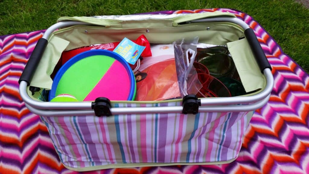 A striped basket filled with colourful outdoor toys and items sits on a vibrant patterned blanket on grass. Visible items include a neon green and pink paddle ball set, frisbees, and packaged goods, ready for a picnic or outdoor activity.