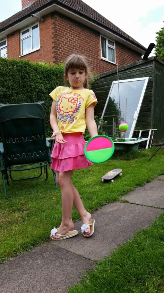 A young girl stands on a garden path, holding a neon green and pink bat. She wears a yellow T-shirt, pink skirt, and sandals with white flowers. Her expression is playful. Garden chairs, a skateboard, and a glass pane are visible in the background.