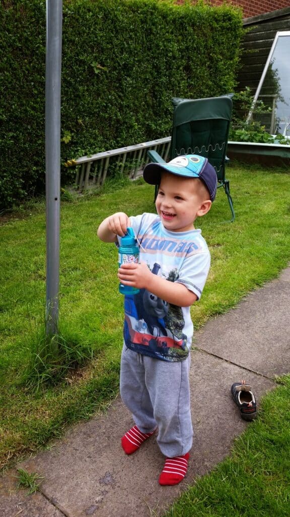 A smiling young boy in a Cars-themed cap and T-shirt stands on a garden path, holding a blue drinks bottle. He wears grey tracksuit bottoms and red-striped socks. A single shoe lies on the grass nearby; a green chair, ladder, and hedges are in the background.