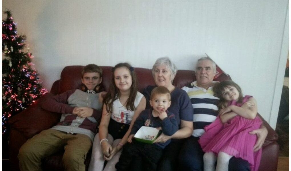 A family of six, including two adults and four children, sit together on a red sofa. A decorated Christmas tree is visible on the left. Everyone is smiling; one girl in a pink dress hugs the older man, while the youngest child sits on an adult’s lap.