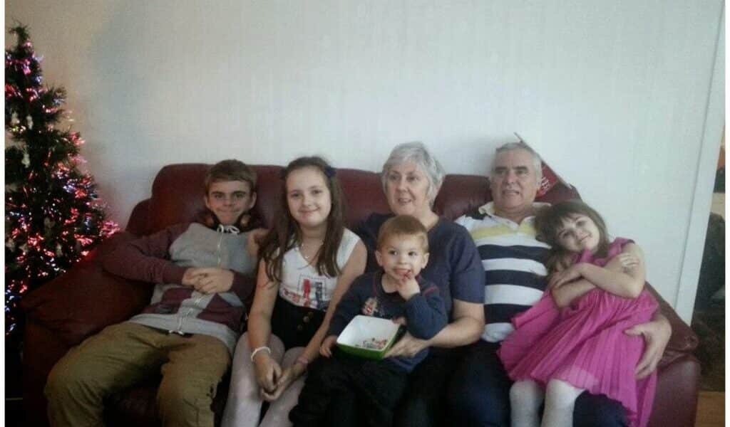 A family of six, including two adults and four children, sit together on a red sofa. A decorated Christmas tree is visible on the left. Everyone is smiling; one girl in a pink dress hugs the older man, while the youngest child sits on an adult’s lap.