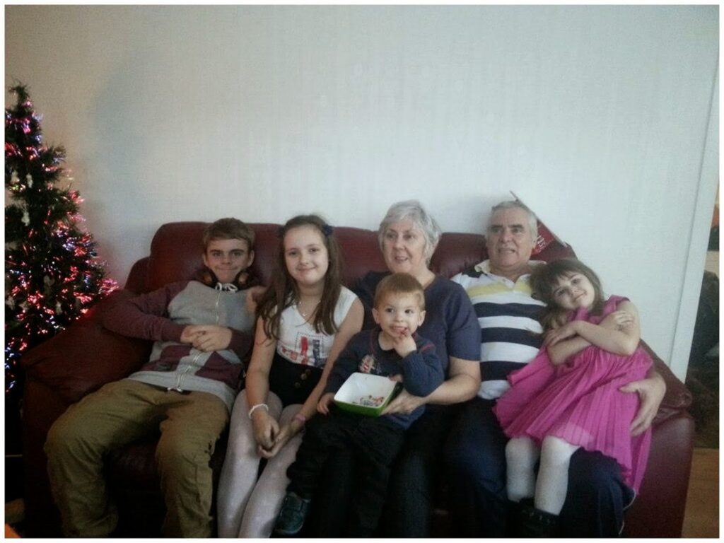 A family of six sits on a red sofa. Two adults, a woman with grey hair and a man in a striped shirt, are surrounded by four children. A Christmas tree with lights is to the left. Everyone is smiling or relaxed, with the youngest girl wearing a bright pink dress.