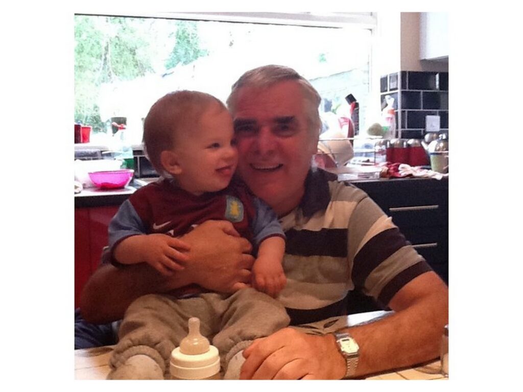 An older man with grey hair, wearing a striped shirt, smiles whilst holding a laughing toddler on his lap in a kitchen. The toddler has fair hair and wears a maroon and blue shirt. A baby bottle sits on the table in front of them.