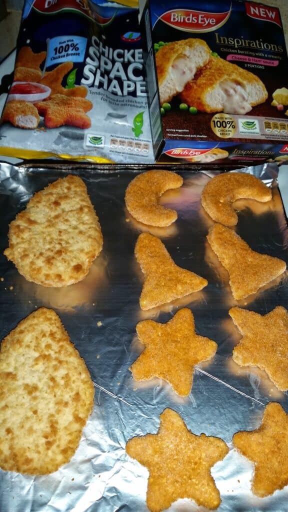 Two opened boxes of Birds Eye chicken nuggets are shown above a baking tray lined with foil. On the tray are shaped nuggets: two are teardrops, one is a crescent moon, one is a star, and one is a witch’s hat. The nuggets are golden-brown.