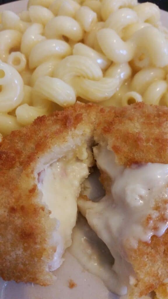 A close-up of creamy macaroni cheese next to a breaded, golden-brown chicken burger split open, revealing melted cheese flowing from the centre. The food is served on a white plate.