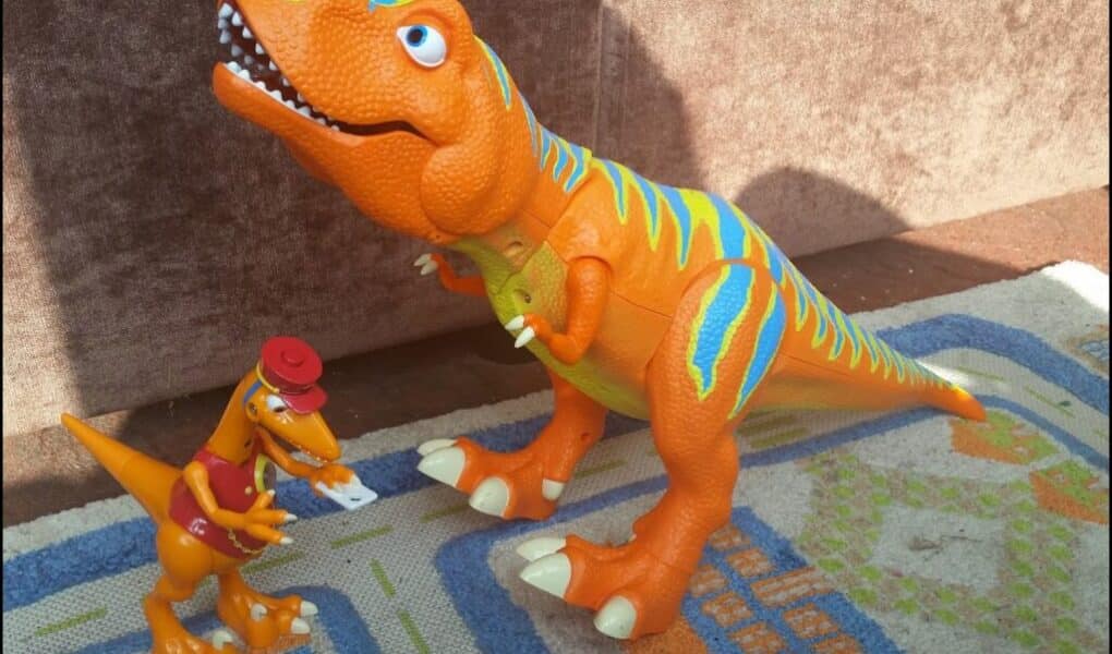 Two dinosaur toys on a patterned rug: a large orange T-Rex with blue and yellow stripes, standing with its mouth open, and a smaller orange dinosaur wearing a red hat and shirt, holding a piece of paper. Sunlight casts shadows behind them.