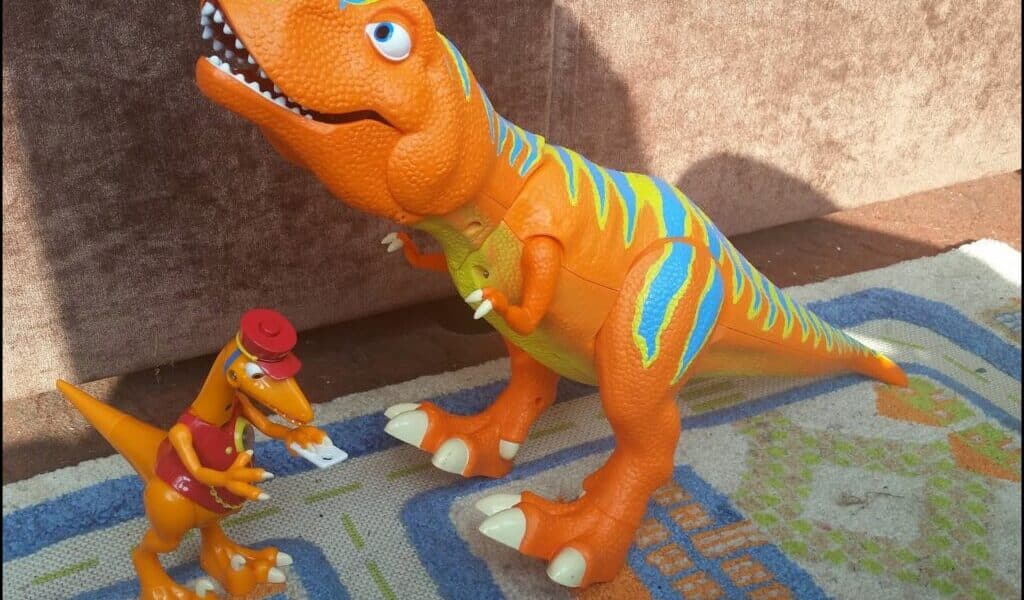 Two dinosaur toys on a patterned rug: a large orange T-Rex with blue and yellow stripes, standing with its mouth open, and a smaller orange dinosaur wearing a red hat and shirt, holding a piece of paper. Sunlight casts shadows behind them.
