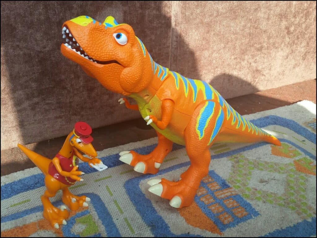 Two orange toy dinosaurs stand on a patterned carpet. The larger dinosaur has blue and green stripes, big eyes, and an open mouth with visible teeth. The smaller dinosaur wears a red hat, holds a letter, and has a postbag over its shoulder.