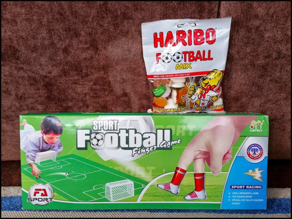 A bag of Haribo Football Mix gummies sits on a brown sofa behind a green box labelled “Sport Football Finger Game.” The box shows a child playing table football using fingers with illustrated boots, and various sport-themed images and logos.