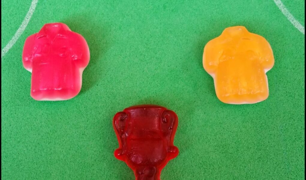 Three gummy sweets on a green surface with white lines resembling a football pitch: a pink shirt, a yellow shirt, and a red trophy-shaped sweet centred below them, suggesting a football theme or competition.