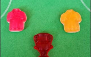 Three gummy sweets on a green surface with white lines resembling a football pitch: a pink shirt, a yellow shirt, and a red trophy-shaped sweet centred below them, suggesting a football theme or competition.