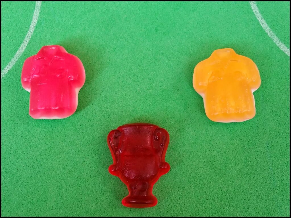 Three gummy sweets are on a green surface with white lines resembling a football pitch. Two gummies are shaped like shirts, one pink (left) and one yellow (right), and one gummy in the centre is shaped like a trophy and is a dark red colour.