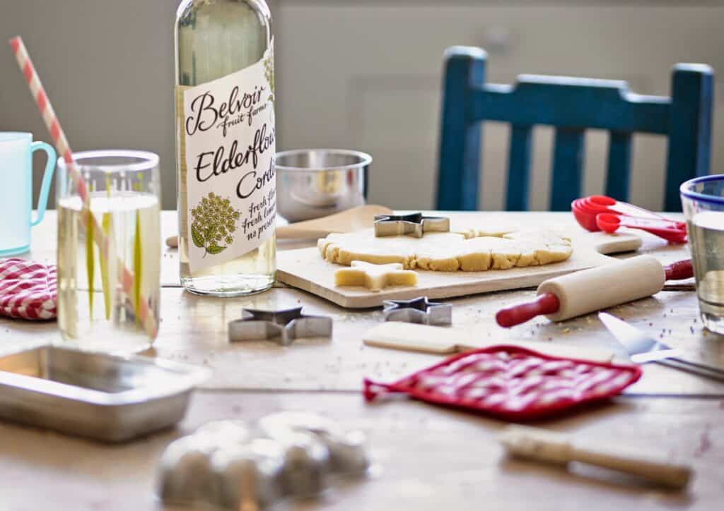 A wooden table with baking supplies: a rolling pin, biscuit cutters, dough, a bottle labelled "Belvoir Elderflower," a red oven glove, a metal tin, a glass with a straw, measuring cups, and a blue chair in the background. The scene is bright and cosy.