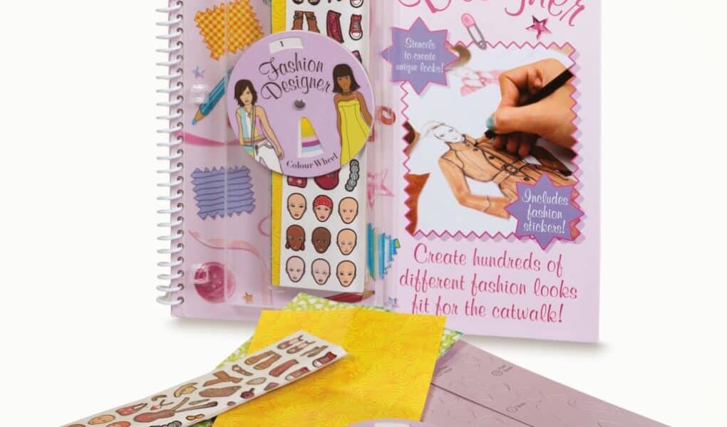 A spiral-bound “Fashion Designer” activity book with drawing tools, sticker sheets showing faces, clothes, and accessories, a coloured pencil set, patterned papers, and an illustrated cover showing a hand sketching a fashion design with styled text and vibrant colours.