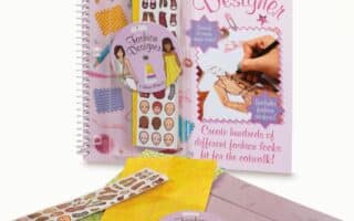 A spiral-bound “Fashion Designer” activity book with drawing tools, sticker sheets showing faces, clothes, and accessories, a coloured pencil set, patterned papers, and an illustrated cover showing a hand sketching a fashion design with styled text and vibrant colours.