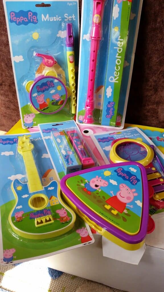 A variety of colourful Peppa Pig-themed toy musical instruments in packaging, including a recorder, drum, guitar, triangle, and toy harmonica. Each package features illustrations of Peppa Pig and friends on a green field with blue sky backgrounds.