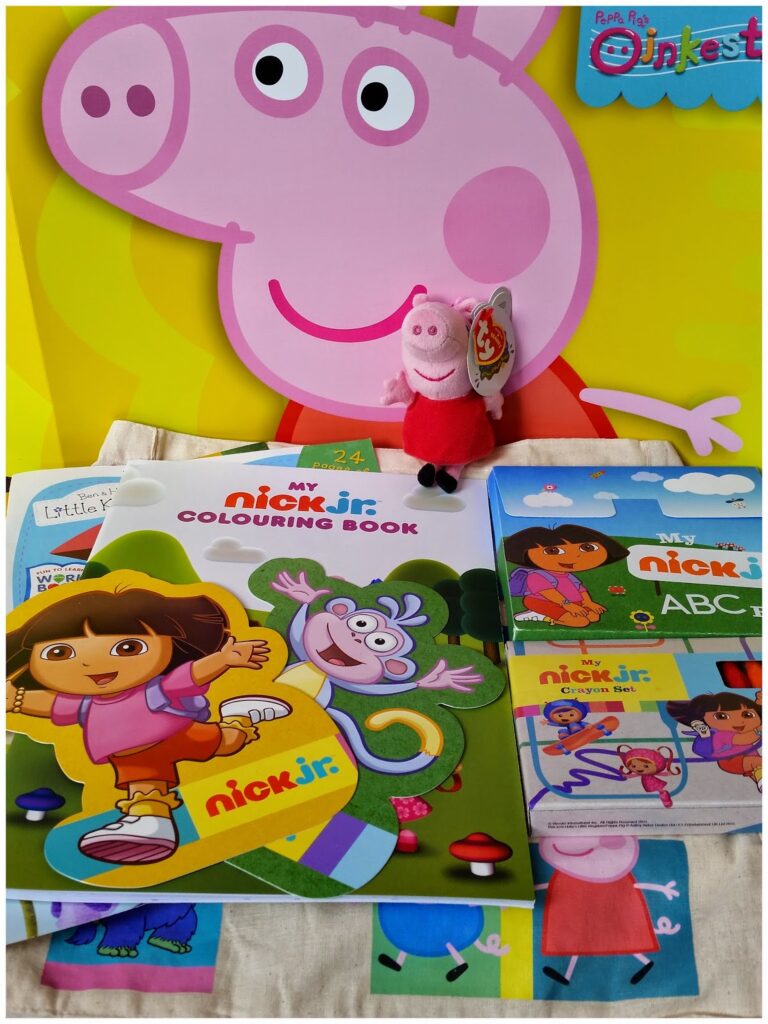 A colourful display featuring a large Peppa Pig poster, a small Peppa Pig soft toy, a Nick Jr. colouring book with Dora and Boots on the cover, Dora the Explorer ABC flashcards, and other children's activity items arranged on a table.