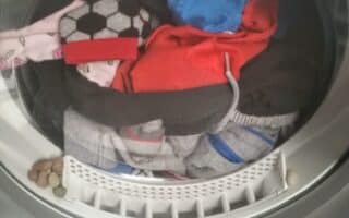 A front-loading washing machine filled with colourful clothes, including a football-patterned sock and blue shorts. Several pieces of dry pet food are trapped in the rubber rim around the drum’s opening, just below the door.