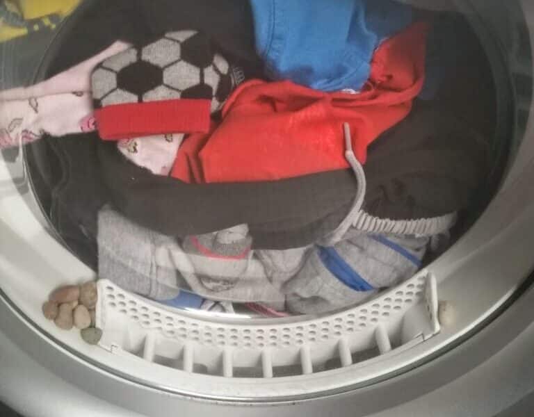 A front-loading washing machine filled with colourful clothes, including a football-patterned sock and blue shorts. Several pieces of dry pet food are trapped in the rubber rim around the drum’s opening, just below the door.