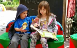 Two children sit on a colourful bench outdoors. The older girl reads a large book while the younger boy, in a blue hoodie, gestures with his hand. Behind them is a chalkboard with "Ameen" written on it and lush green plants in the background.