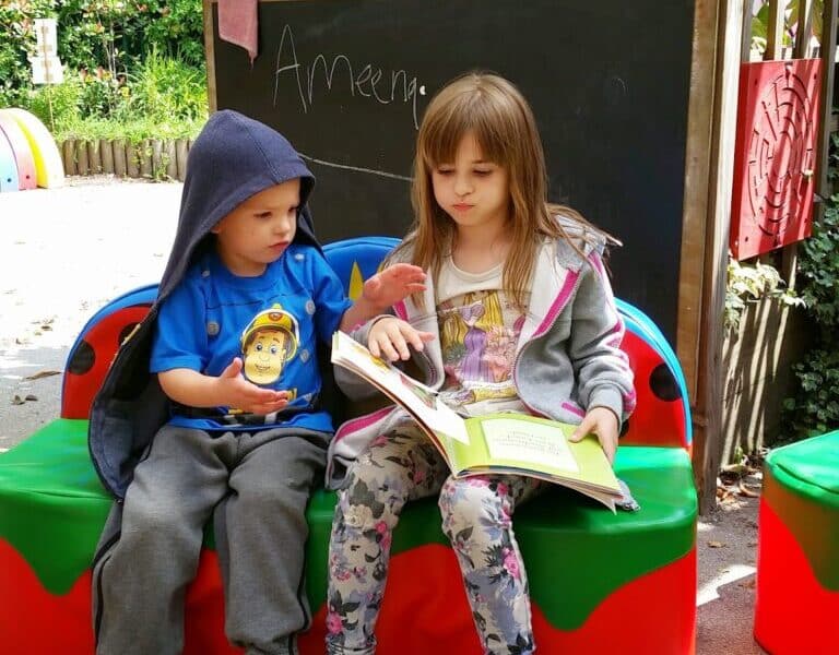 Two children sit on a colourful bench outdoors. The older girl reads a large book while the younger boy, in a blue hoodie, gestures with his hand. Behind them is a chalkboard with "Ameen" written on it and lush green plants in the background.