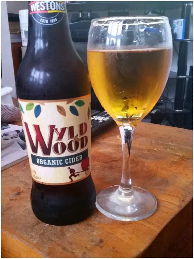 A bottle of Westons Wyld Wood Organic Cider sits on a wooden table next to a filled wine glass of golden cider. In the background, blurred objects like a TV remote, papers, and part of a sofa are visible.