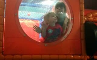 Two young children are inside an indoor play structure, looking and pressing their faces and hands against a clear, round window set in an orange wall. The background is bright and colourful, with play equipment visible behind them.