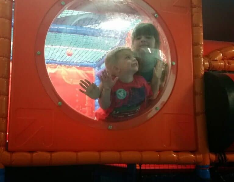 Two young children are inside an indoor play structure, looking and pressing their faces and hands against a clear, round window set in an orange wall. The background is bright and colourful, with play equipment visible behind them.