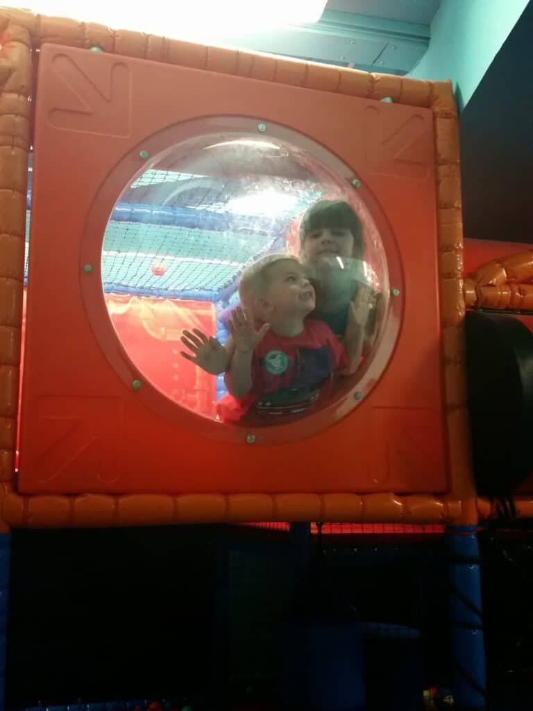 Two young children are inside an indoor play structure, looking out through a clear, circular bubble window set in an orange frame. One child is smiling, and both have their hands pressed against the bubble, with colourful play equipment visible around them.