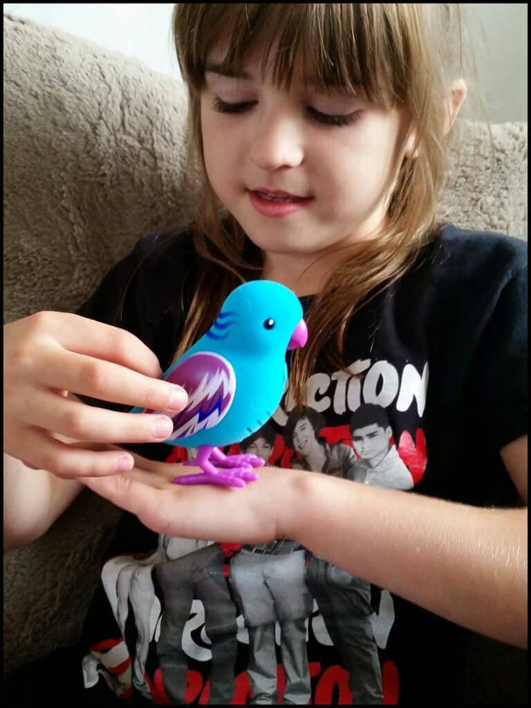 A young girl with light brown hair and fringe smiles while holding a blue and purple toy bird in her hands. She wears a black T-shirt with black and white photos of people and red text on it, sitting on a beige, textured sofa.