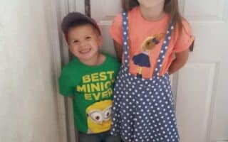 A young boy wearing a green “Best Minion Ever” T-shirt, blue shorts, socks, trainers, and a red cap stands smiling beside a girl in a polka dot pinafore skirt and orange Minion T-shirt. They pose indoors in front of a closed white door.