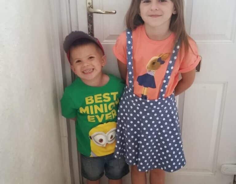 A young boy wearing a green “Best Minion Ever” T-shirt, blue shorts, socks, trainers, and a red cap stands smiling beside a girl in a polka dot pinafore skirt and orange Minion T-shirt. They pose indoors in front of a closed white door.