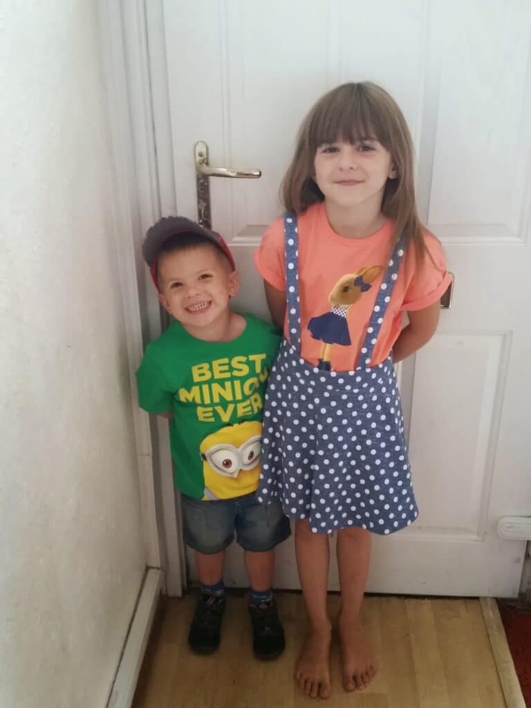 A smiling young boy in a green “Best Minion Ever” shirt, denim shorts, a cap, and dark shoes stands next to a barefoot girl in a peach T-shirt and blue polka-dot pinafore skirt. They pose together by a white door indoors.