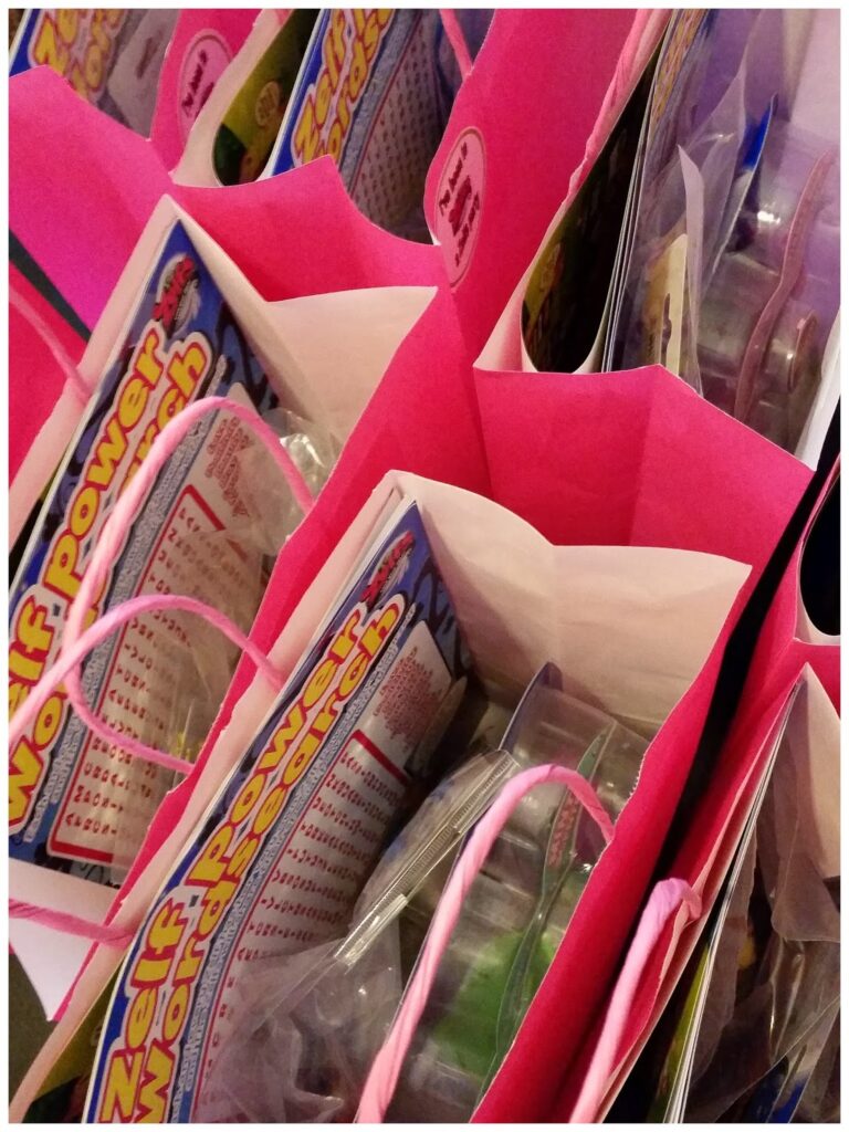 Several pink and white gift bags stand side by side, viewed from above. Each bag is filled with colourful Zelf Power Watch packaging and other small toys or treats, visible just above the edge of the bags. Pink twisted handles are draped over the sides.