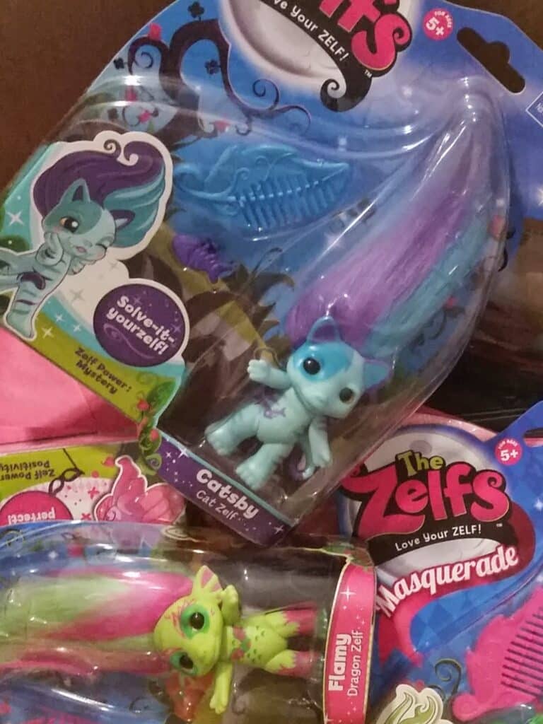 Two colourful toy dolls from "The Zelfs" series in plastic packaging. One is blue with purple hair ("Catzby Cat Zelf") and the other is green with pink hair ("Flamy Dragon Zelf"). Accessories and decorative designs are visible in the packages.