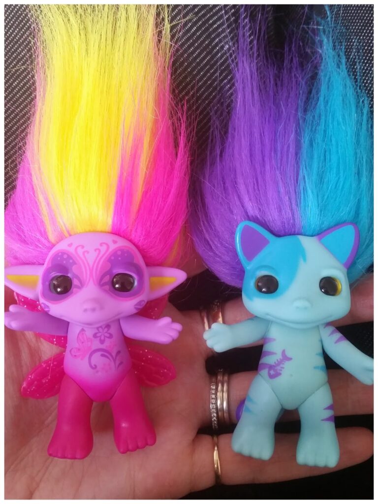 Two small toy dolls are held in a person's hand. The left doll has bright pink skin, large eyes, yellow and pink hair, elf-like ears, and wing designs. The right doll is blue with turquoise stripes, cat-like ears, a purple tail, and bright purple hair.