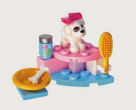 A white toy dog with a pink bow sits on pink and blue building bricks. In front, a yellow brush stands upright, a yellow bowl with a white bone rests nearby, and a tin with a paw print design is also present, resembling a pet grooming set-up.