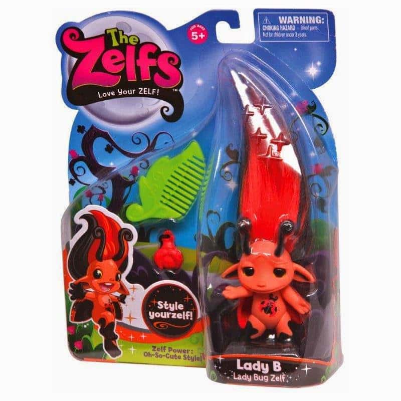 A packaged “The Zelfs” toy featuring Lady B, a red doll with black accents and tall, spiky hair decorated with ladybird spots. The set includes a green comb and a red charm. Packaging has colourful graphics and a “Style yourself!” slogan. For ages 5+.