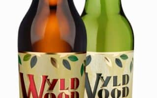 Two bottles of Westons Wyld Wood cider stand side by side. The brown bottle is labelled “Organic Cider” with a red apple graphic, whilst the green bottle is labelled “Organic Pear Cider” with a pear graphic. Both labels feature leaves and a small tree silhouette.