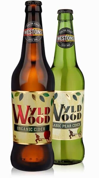Two bottles of Westons Wyld Wood cider stand side by side. The brown bottle is labelled “Organic Cider” with a red apple graphic, whilst the green bottle is labelled “Organic Pear Cider” with a pear graphic. Both labels feature leaves and a small tree silhouette.