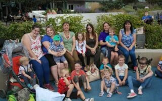 A group of adults and children sit closely together outdoors, smiling at the camera. Some adults hold young children on their laps. There are pushchairs, bags, and greenery in the background, suggesting a family outing or gathering in a park-like setting.