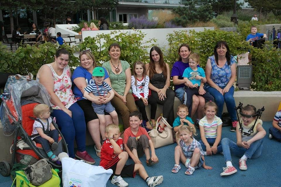 A group of adults and children sit closely together outdoors, smiling at the camera. Some adults hold young children on their laps. There are pushchairs, bags, and greenery in the background, suggesting a family outing or gathering in a park-like setting.
