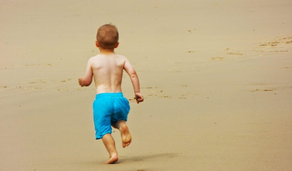 A young child with short hair, wearing blue shorts and no shirt, runs barefoot on a sandy beach. The child is facing away from the camera, with arms slightly bent and footprints visible in the smooth, golden sand. The background is empty beach.