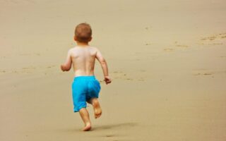 A young child with short hair, wearing blue shorts and no shirt, runs barefoot on a sandy beach. The child is facing away from the camera, with arms slightly bent and footprints visible in the smooth, golden sand. The background is empty beach.