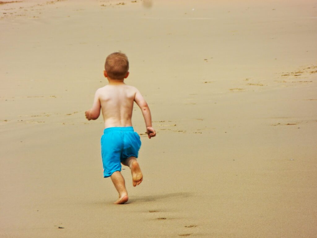 A small child with short light brown hair, wearing bright blue shorts and no shirt, is running barefoot on a sandy beach. The child is seen from behind, and the beach appears empty, with soft, smooth sand stretching ahead.
