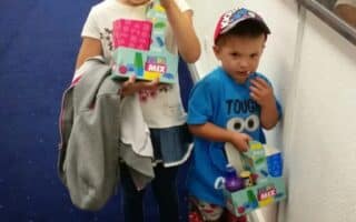 A young girl smiles and gives a thumbs-up while holding a drink and box of sweets. Beside her, a younger boy in a blue shirt and hat holds a similar box, looking shy. A tub of popcorn sits on the floor. They stand in a corridor with film posters.