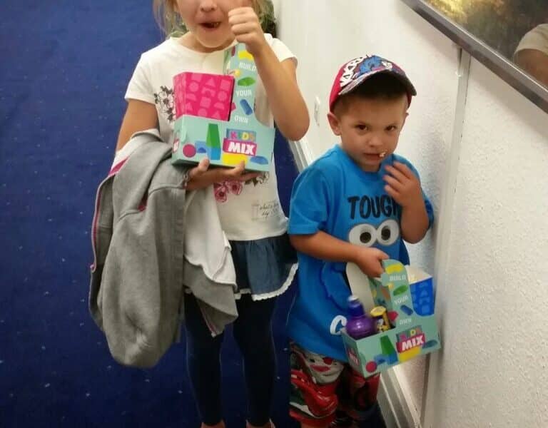 A young girl smiles and gives a thumbs-up while holding a drink and box of sweets. Beside her, a younger boy in a blue shirt and hat holds a similar box, looking shy. A tub of popcorn sits on the floor. They stand in a corridor with film posters.